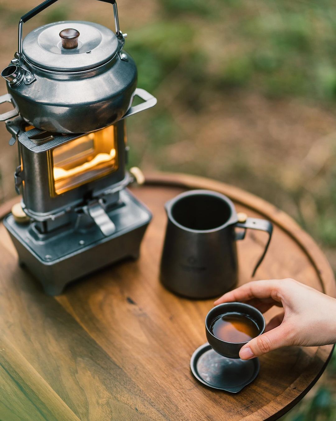 vintage oil lamp with a silver tea kettle on top setting on a wooden table outside next to a woman drinking tea