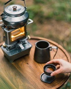 vintage oil lamp with a silver tea kettle on top setting on a wooden table outside next to a woman drinking tea